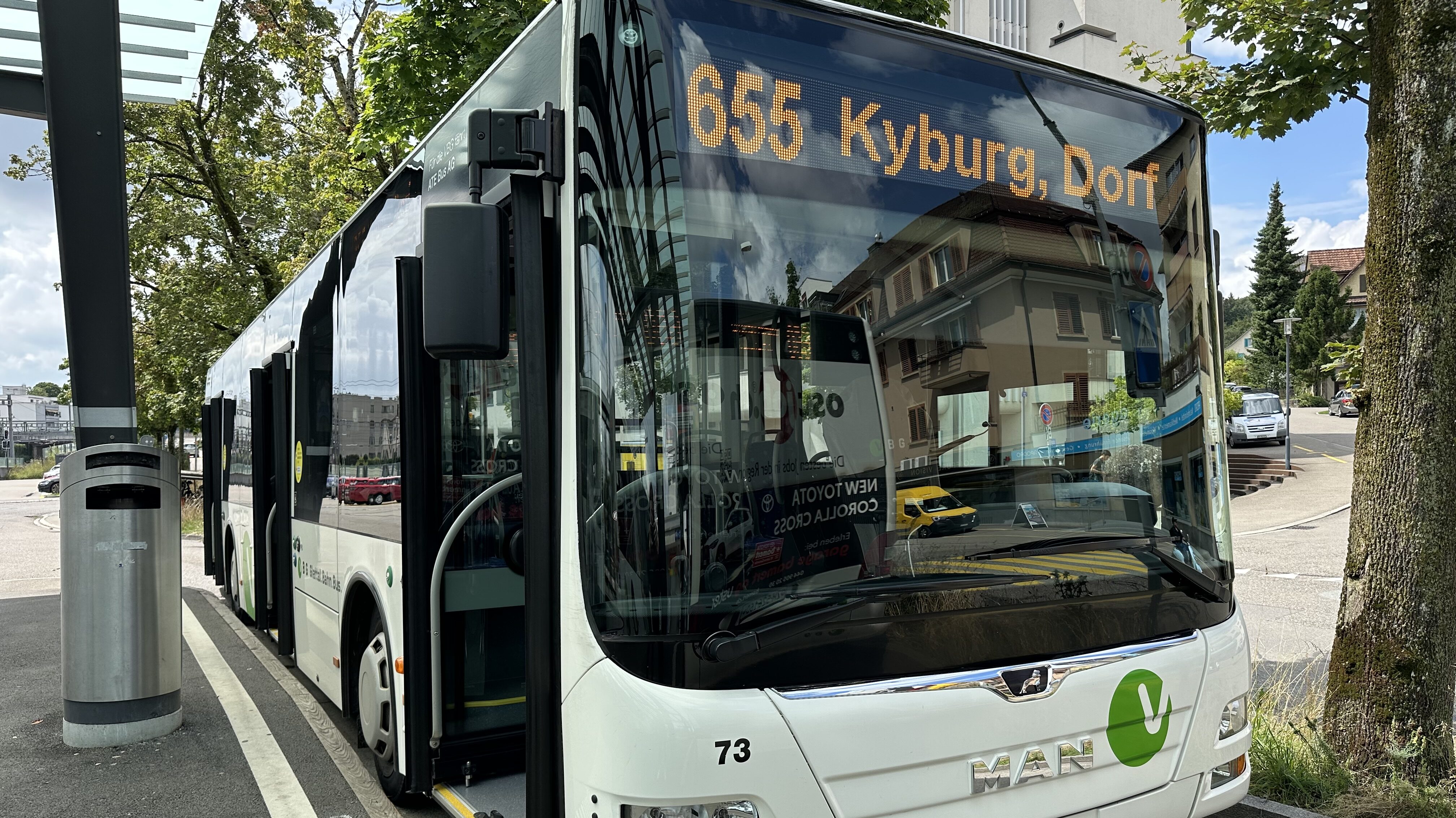 Ein VBG-Bus steht an der Bushaltestelle, dahinter blühen die Bäume im sommerlichen Kleid.